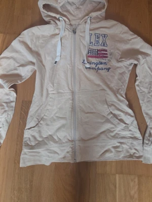 Lexington hoodie - Säljer en beige hoodie med dragkedja och huva. Framtill finns tryck med texten lex och en amerikansk flagga samt blå brodyr. Hoodien har fickor på sidorna och vita snören i huvan. Perfekt till jeans för en avslappnad look.