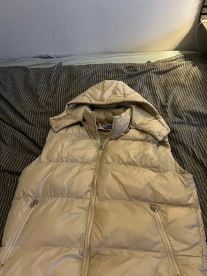 Beige dunväst med huva - Snygg beige dunväst med avtagbar huva och dragkedja framtill. Västen har två fickor med dragkedja och quiltad design. Perfekt för lager-på-lager och ger en clean look till din outfit. Finns lite fläckar