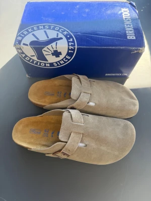Beige mockatofflor från Birkenstock - Säljer ett par snygga beige tofflor från Birkenstock i mjuk mocka. Modellen har en bred rem med justerbart spänne och klassisk korksula för extra komfort. Perfekta för chill dagar hemma eller när du vill ha en avslappnad stil.
