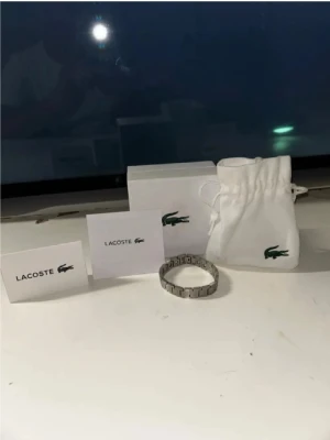  armband från Lacoste - Snyggt armband i silverfärg från Lacoste med stilren, länkad design. Kommer med originalpåse och box, perfekt för dig som gillar klassiska accessoarer med en sportig touch.