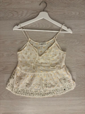 Abercrombie & Fitch guld prickig linnetopp - Supersöt ljusgult linnetopp från Abercrombie & Fitch i vitt med guldiga prickar och broderade blommor. Toppen har tunna axelband, v-ringning med knyt och fina detaljer😊Perfekt för sommarens alla tillfällen! Strl: XS