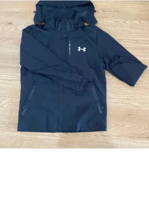Svart vindjacka från Under Armour - Snygg svart vindjacka från Under Armour med vit logga på bröstet och texten 'EXPLORE' vid dragkedjan. Jackan har huva, två dragkedjeförsedda fickor och elastiska muddar. Perfekt för blåsiga dagar och outdoor-aktiviteter.