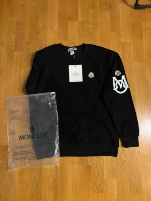 Svart sweatshirt från Moncler - Svart sweatshirt från Moncler med rund halsringning och ribbade muddar. Tröjan har Moncler-logga på bröstet och ett stort vitt M-tryck på ena ärmen. Perfekt för dig som gillar streetwear och exklusiva märken. Passar S ganska tight på M