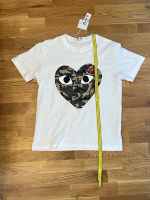 Vit t-shirt med camo-hjärta från Comme des Garçons - Säljer en vit t-shirt från Comme des Garçons Play med ett stort hjärta i camouflage-mönster och en liten röd hjärtdetalj på bröstet. Klassisk rund hals och korta ärmar. Perfekt statement-plagg för dig som gillar streetwear.