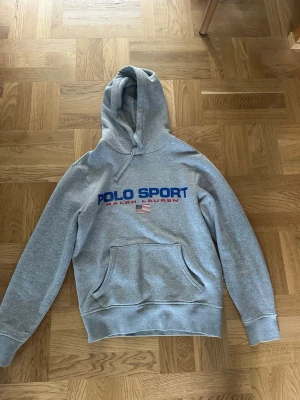 Grå hoodie Polo Sport Ralph Lauren - Snygg grå hoodie från Polo Sport Ralph Lauren med blå och röd logga på bröstet samt amerikansk flagga. Klassisk känguruficka framtill och justerbar huva. Perfekt för en avslappnad och sportig stil.