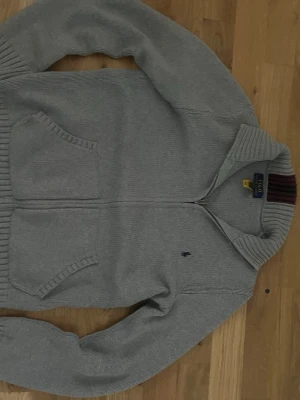 Grå ziptröja från Polo Ralph Lauren - Snygg grå ziptröja från Polo Ralph Lauren med två stora fickor framtill och ribbade muddar. Tröjan har en liten broderad logga på bröstet och en krage med randiga detaljer på insidan. Perfekt för en chill och stilren look.