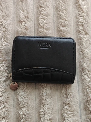 Svart plånbok från Wera - Svart plånbok från Wera med dragkedja runtom och präglad detalj i nederkant. Plånboken är i slätt konstläder med en liten metallbricka på dragkedjan där det står Wera. Perfekt storlek för kort och kontanter.