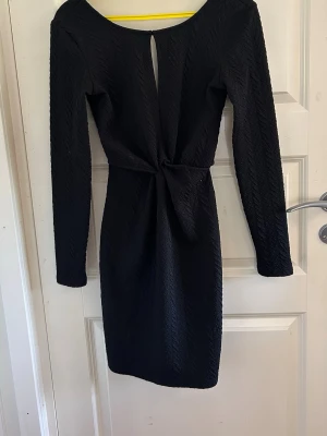 Svart långärmad kortklänning - Snygg svart kortklänning med lång ärm och twistad detalj i midjan. Klänningen har en liten öppning framtill vid halsen och är tillverkad i ett strukturerat tyg som ger en cool look. Perfekt för dig som gillar stilrena plagg med det lilla extra.