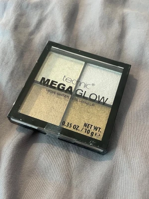 Technic Mega Glow Highlighter - Säljer en Technic Mega Glow highlighterpalett med fyra skimrande nyanser i guld, champagne och silver. Perfekt för att skapa en glowig look. Kommer i en fyrkantig plastförpackning med genomskinligt lock. Oöppnad present