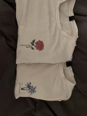 Vita t-shirts med tryck - Två vita t-shirts med coola tryck framtill. Ena har en röd ros med svart text, den andra har palmer och texten 'Summer'. Båda har rund hals och korta ärmar, perfekta för en avslappnad stil.