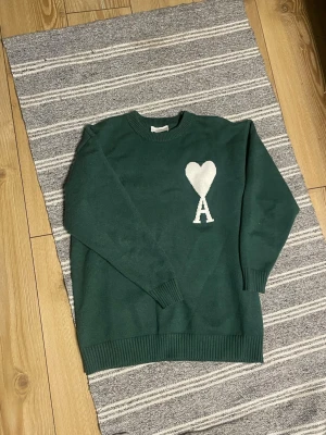 Grön stickad tröja från Ami - Mörkgrön stickad tröja från Ami Alexandre Mattiussi med vit hjärtlogga och bokstaven A på bröstet. Tröjan har rund halsringning, ribbade muddar och är gjord i mjukt material. Perfekt för dig som gillar stilrena detaljer.