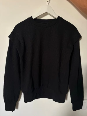 Svart sweatshirt från Ateks - En stilren svart sweatshirt från Ateks med rund halsringning och ribbade muddar vid ärmslut och nederkant. Tröjan har en enkel och clean design utan tryck eller huva, perfekt för en avslappnad look. Materialet ser ut att vara mjukt och bekvämt.