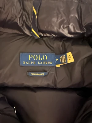 Svart pufferjacka från Polo Ralph Lauren - Snygg svart pufferjacka från Polo Ralph Lauren med glansig finish och röd logga på bröstet. Jackan har huva, flera dragkedjor och quiltad design som ger en cool och sportig vibe. Perfekt för kalla dagar när du vill hålla stilen.