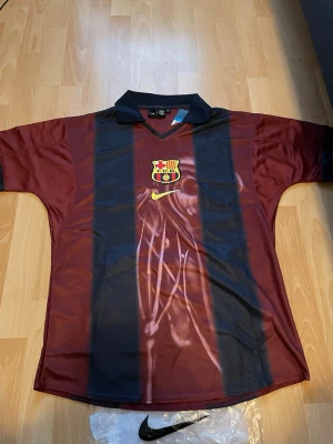 Barcelona x Cactus Jack matchtröja - Unik Barcelona matchtröja i vinrött och marinblått med klubbmärke och Nike-logga på framsidan. Baksidan har 'Cactus Jack', nummer 2 och 'BARÇA' tryckt. Klassisk krage och korta ärmar. Perfekt för dig som älskar fotboll och streetwear.