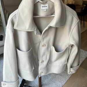 Kort kappa -  Kort jacka i en rak och vid modell med krage, knappar framtill och stora oversize fickor på framsidan. Jackan har även ett band vid ärmslut med en knapp som en snygg detalj.Använd fåtal gånger så i väldigt fint skick. Nypris 899kr💞 köparen står för frakt