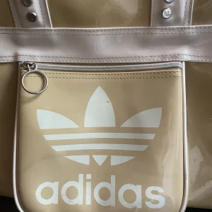 Adidas väska - Adidas väska i lackacktigt material? Ficka på framsidan och mycket rymlig. Finns en platta i botten så den håller formen samt stabilitet i väskan. 