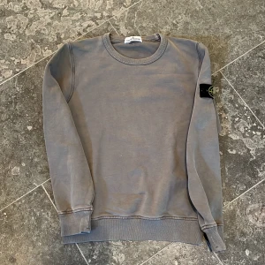 Stone Island tröja junior  - Säljer en grågrön Stone Island tröja. Mkt fint skick förutom ett mindre hål på ryggen som enkelt kan lagas, se bild. Storlek 10, ca 145