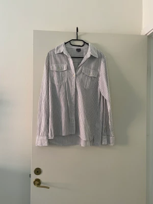 Vit blårandig skjorta  - Tunnt blårandig vit bomullsskjorta med v-ringning. Ärmarna går att vika upp och fästa med knapp. 