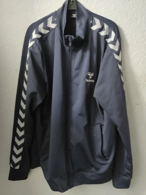 Träningsjacka Hummel XL - En snygg träningsjacka i blått med gråa ränder på ärmarna. Den är i ett väldigt gott skick.