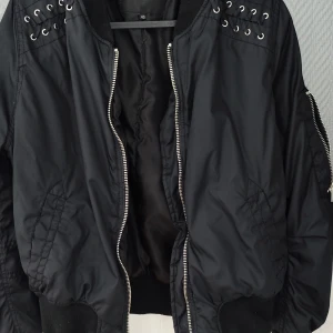 Bomberjacka - Svart bomberjacka med snörningsdetaljer på rygg och fram vid axlarna. Sparsamt använd storlek 40 
