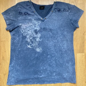 Vintage G-Star T-Shirt - En extremt snygg ljusblå tröja med ännu snyggare detaljer! Ett tryck på framsidan likt Affliction och volangliknande axlar, samt fungerande knappar längs tröjans krage! Väldigt skönt material, perfekt för sommaren! Står storlek 4, sitter som en M