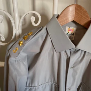 Svensk militärskjorta – vintage – 50–60-tal – suveränt skick - Finish Charmeuse Nylon. Made in Sweden.  Grå färg. Axelklaffar med tre guldstjärnor (kaptensbeteckning, tror jag). Och siffran 3 i guld.  Storlek: 39 (M).  Bröst: 56 cm. Längd: 85 cm.