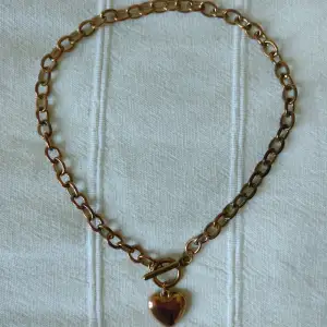 Halsband i metall med berlock i form av ett hjärta. Ca 40 cm långt. Mässings-/kopparfärgat - färgen skiftar lite. Kan skickas med ett frimärke (18 kr).