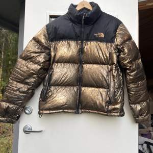 Limited edition dunjacka i bräcklig guld färg från The North Face. Logan baktill har tappat några bokstäver och färgen har börjat slitas bort på vissa ställen. Något liten i storleken.