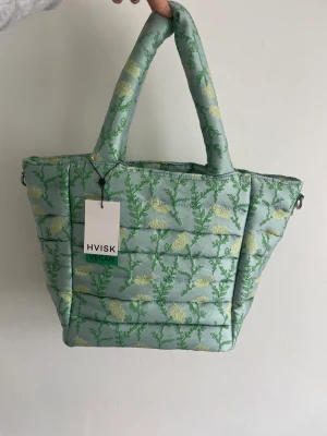 Hvisk väska - En trendig väska från köpenhamnsmärket Hvisk. Blått material med gröna och gula blommönster. Prislappen sitter kvar, aldrig använd. Kommer även med en avtagbar rem som gör den till crossbody. Får plats med en Macbook.  H1805 Valley Medium Yarrow 014 Blue 