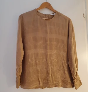 Glansig blus  - Glansig skjortblus med fladdermusärm,  storlek 36 Sandfärgad blus med fladdermusärm/pinguinärm från Yaha. Knappar på baksidan.  Knappt använd. Mycket bra skick. Inga fläckar eller skador. Längd: 65 cm