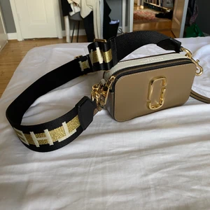 Marc Jacobs väska - Köpte denna bruna Marc Jacobs väska i köpcentrumet KaDeWe i Berlin för 2 år sedan, men knappt använt. Så väskan är i väldigt bra skick. Väskan är brun och vit på sidorna, guldiga detaljer, guldig och svart band till väskan. Köpte den för 3200 kr.