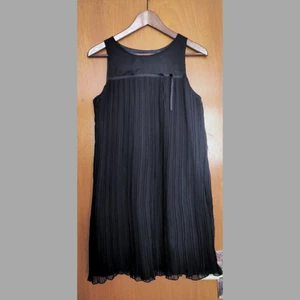 Svart 20tals-inspirerad klänning - Den här svarta plisserade klänningen fullkomligt andas 20-tal och storartade Gatsby-fester! Den är fodrad i satin på insidan och med lätt draperande chiffong på utsidan. Den är i en tidsenligt utsvängd modell och har även ett band vid bysten med en dekorativ rosett fram. Mycket bekväm och verkligen en pärla!  Finns en liten fläck i ena hörnet av plisseringen, som ej syns om man inte viker upp tyget. Tycker ej det märks när klänningen är på överhuvudtaget. Avhämtning i Stockholm eller skickas!