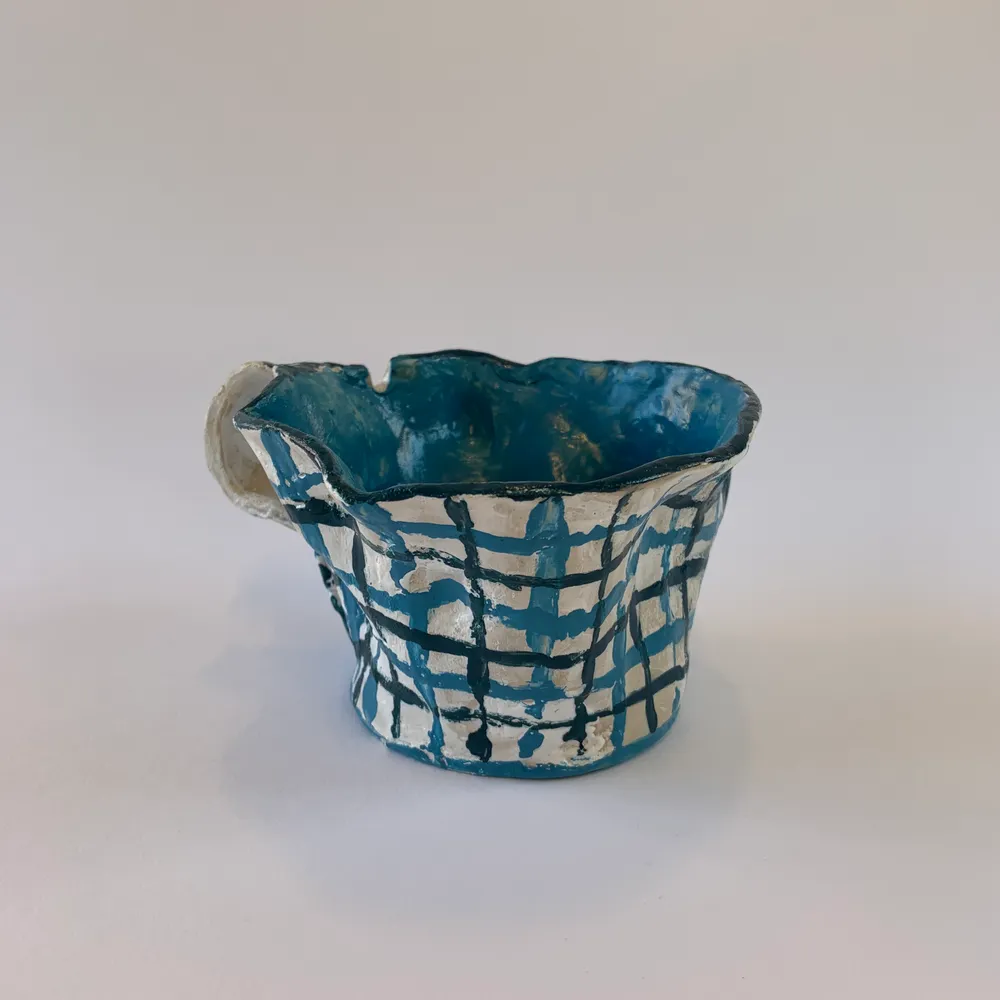 En handgjord kopp i keramik. Koppens yttre har en vit bakgrund med blåa rutor, koppen har även en blå insida. I koppen finns en skåra som är perfekt utformad för bland annat en sked eller tepåse. . Övrigt.