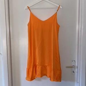 Orange kortklänningen i sidenlikande tyg - Kort klänning, har tunna band med silvriga spännen samt en volang längst ner. 