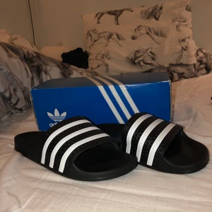 Adidas Tofflor Adilette  - Svarta Adidas tofflor i storlek 39. Box medföljer inte vid frakt, endast vid upphämtning. DM för vidare info! Använda FÅTAL gånger, dvs nyskick. Originalpriset är 379kr. Köparen står för frakt! 💕