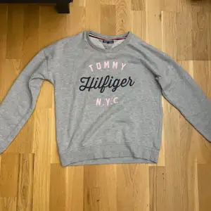 En grå sweatshirt med blå/rosa text. En tommy Hilfiger tröja. Använd ett par gånger för ca 2/3 år sen. Skön att ha på sig. 