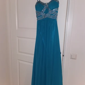 Festklänning - Ljusblå långklänning med detaljer på framsidan. I använt skick, har däremot ett litet hål längst ner på framsidan varvid priset är anpassat efter defekten. Därutöver är den i god skick