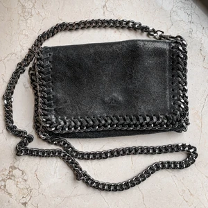Svart Handväska med Silvrig Kedja - Svart Stella McCartney inspirerad axelremsväska med silvrig kedja. Sparsamt använd och i gott skick. Knäppe med blixtlås. Går att ta av kedjan. 