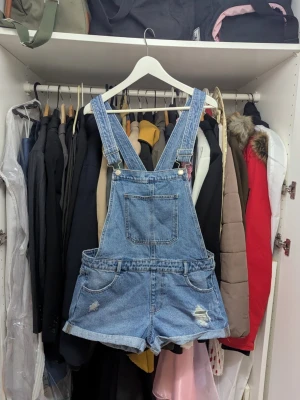 Blå jeansshorts hängselbyxor - Snygga blå jeansshorts hängselbyxor i klassisk denim med slitna detaljer och fransade benslut. Flera fickor fram och bak samt justerbara axelband. Perfekt för en avslappnad och trendig stil.