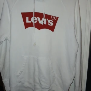 Vit hoodie från Levi's med röd logga - Vit hoodie från Levi's med stor röd logga på bröstet. Klassisk känguruficka framtill och justerbar huva med dragsko. Mjuk bomullskänsla och ribbade muddar vid ärmslut och nederkant. Perfekt för en avslappnad streetstil.