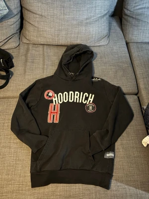 Svart hoodie från Hoodrich med tryck - Säljer en svart hoodie från Hoodrich med stora vita och röda tryck på bröstet och flera patchar. Klassisk känguruficka framtill och ribbade muddar. Hoodien har en huva med dragsko och är perfekt för en streetwear-look.