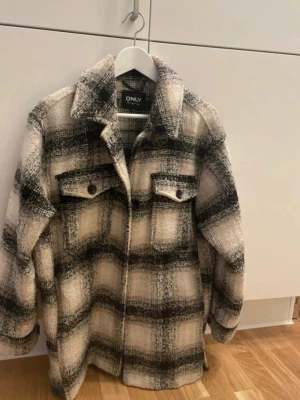 Rutig overshirt från ONLY - Snygg overshirt från ONLY i beige, svart och vitt rutmönster. Jackan har klassisk krage, två bröstfickor med knappar och en rak, avslappnad passform. Perfekt lager-på-lager-plagg med mjukt, borstat material och långa ärmar.