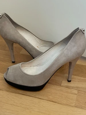 Beige peep-toe pumps Stuart Weitzman - Snygga beige pumps i mocka från Stuart Weitzman med peep-toe och hög klack. Skorna har en svart platåsula och en elegant siluett som ger extra höjd. Perfekta för dig som vill ha en stilren och trendig look.