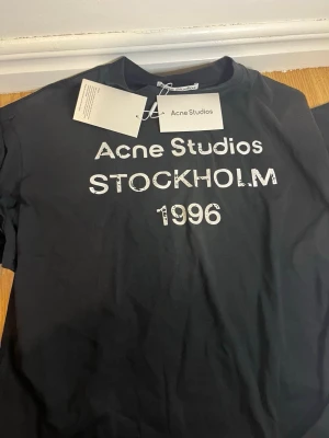 Svart Acne Studios t-shirt med tryck - Cool svart t-shirt från Acne Studios med vit text 'Acne Studios STOCKHOLM 1996' på bröstet. Klassisk passform och rund halsringning, perfekt för en stilren och trendig look.