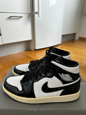 Air Jordan 1 Mid svart/vit (karbonfiber) - Snygga Air Jordan 1 Mid sneakers i svart och vitt med klassisk Nike Swoosh och Air Jordan Wings-logga på sidan. Skorna har ovandel i läder med blanka svarta detaljer och perforerad tå för extra ventilation. Perfekt för dig som gillar streetwear och ikonisk design.