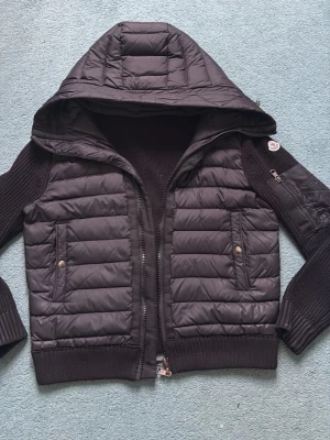Svart pufferjacka från Moncler - Svart pufferjacka från Moncler med stickade ärmar och huva. Jackan har två fickor framtill med knappar och dragkedja. Klassisk Moncler-logga på ärmen och ribbade muddar. Perfekt för kalla vinterdagar.