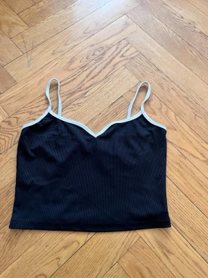 Svart ribbad linnetopp 🌸 - Svart ribbad linnetopp från SHEIN med tunna axelband och vit kant längs urringningen. Toppen har en v-ringad form och är croppad, perfekt för en trendig och enkel stil. Materialet känns mjukt och stretchigt.🌺