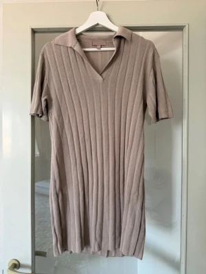 Beige ribbad stickad klänning - Snygg beige stickad klänning med ribbat mönster och kort ärm. Klänningen har en krage och v-ringning som ger en avslappnad men trendig look. Perfekt för dig som gillar stilrena och bekväma plagg. Materialet är mjukt och stretchigt.