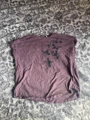 Lila t-shirt med paljettstjärnor - Snygg lila t-shirt med korta ärmar och dekorativa svarta paljettstjärnor på framsidan. Perfekt för dig som vill ha något unikt och glittrigt i garderoben. Mjuk och skön bomullskänsla.