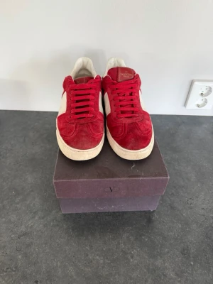 Röda sneakers från Valentino - Snygga röda sneakers från Valentino med vit sidopanel och vita detaljer. Skorna har klassisk snörning och är tillverkade i mocka för en lyxig känsla. Perfekta för dig som vill sticka ut med en färgstark och trendig look.